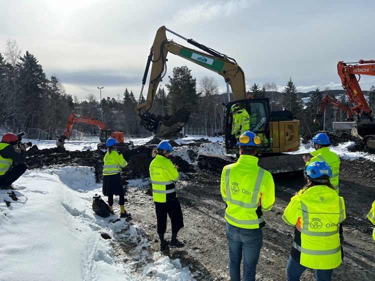 En gruppe mennesker i gule jakker som står i snøen med en gravemaskin